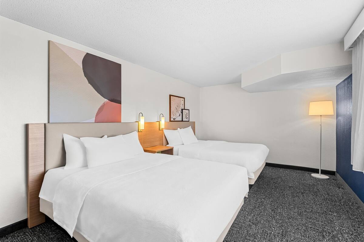 Una habitación de hotel con dos camas, un cuadro abstracto grande, una lámpara de pie y una mesita con una lámpara.