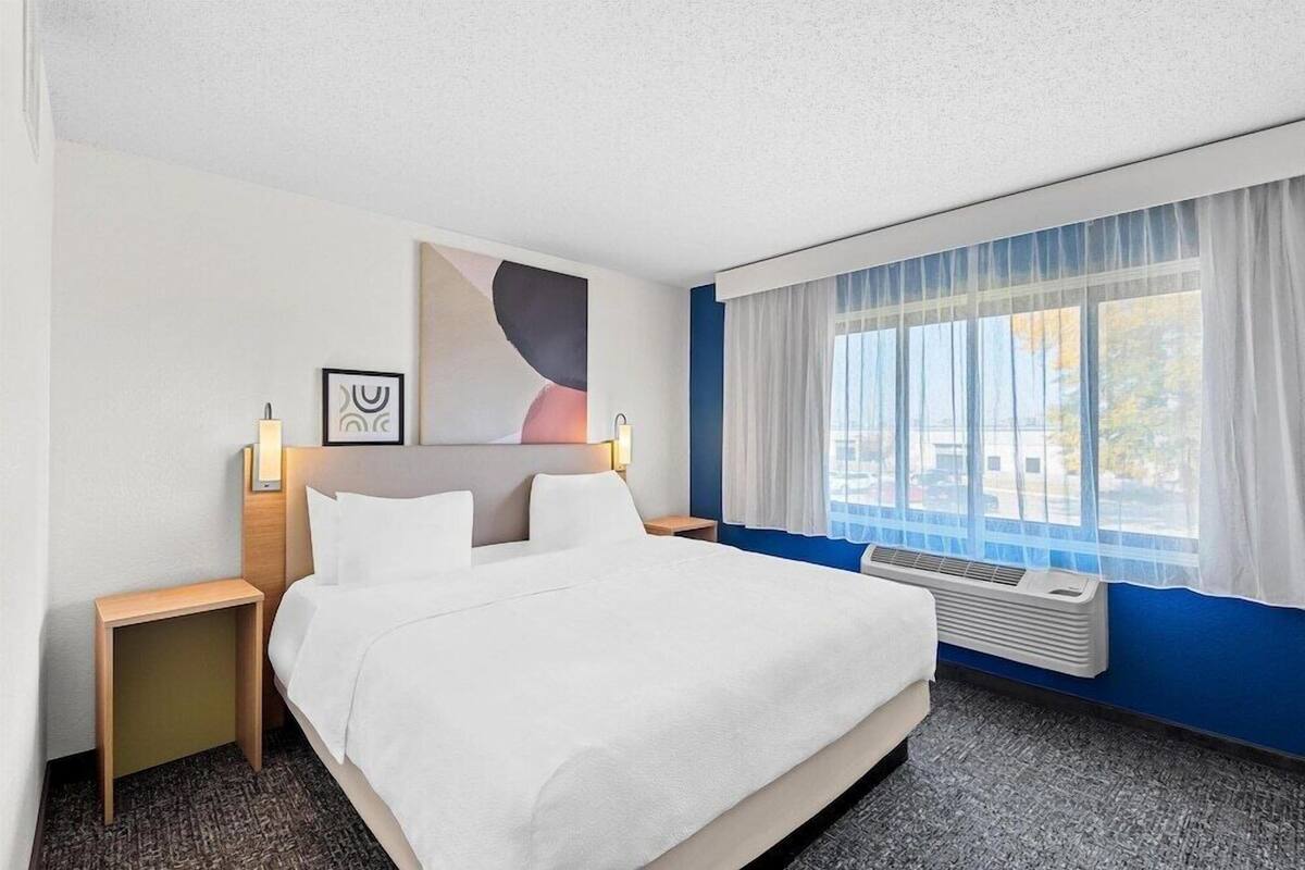 Habitación de hotel con cama, mesitas de noche, una obra de arte colgada en la pared, ventana con cortinas y un equipo de aire acondicionado también montado en la pared.