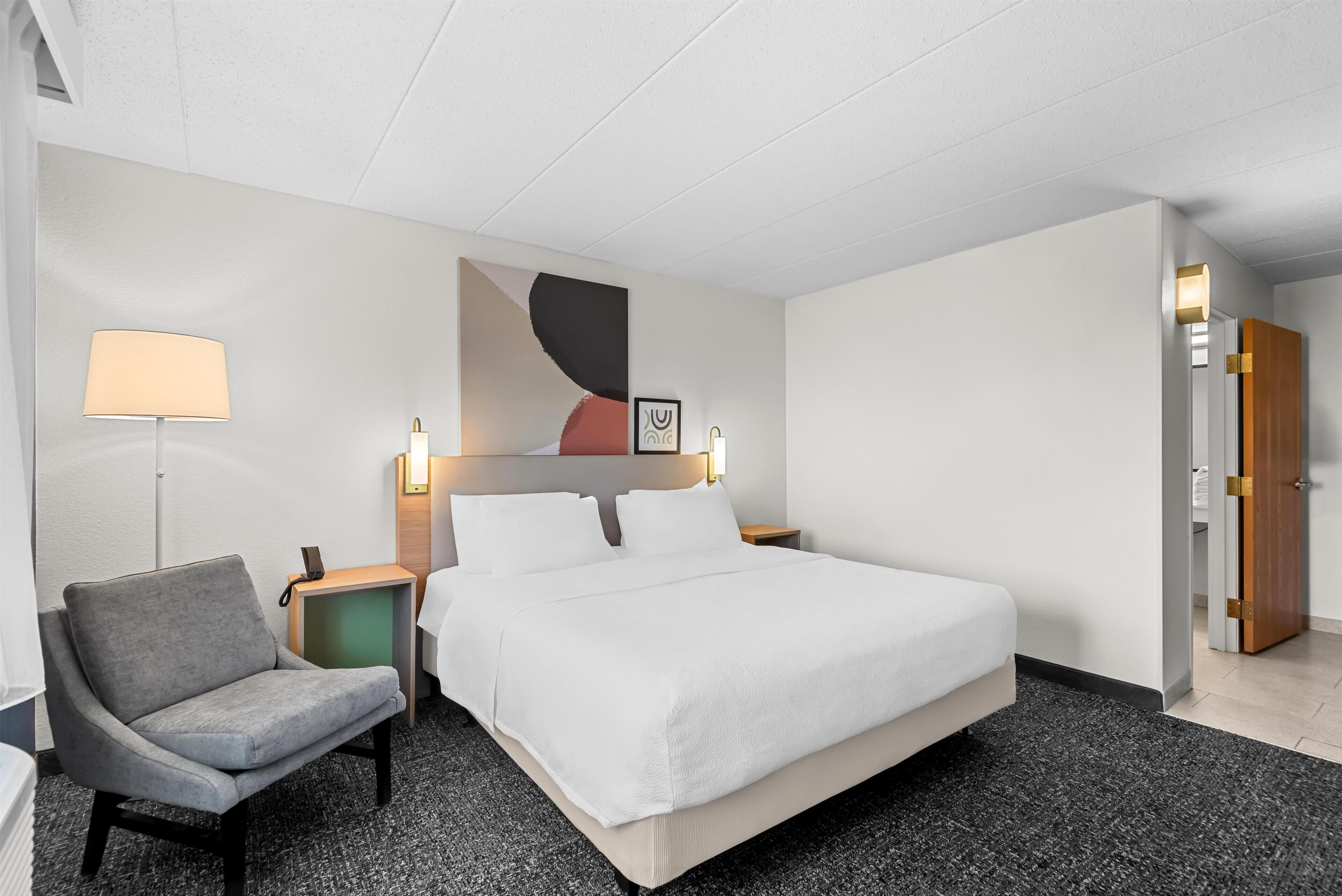 A hotel room with a large bed, a chair, a bedside table with a lamp, and a wall-mounted artwork.