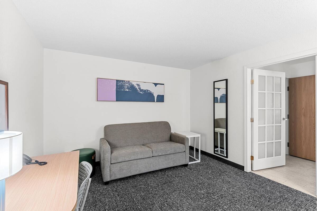 Una habitación moderna con un sofá gris, una mesita auxiliar blanca, un cuadro abstracto enmarcado y una puerta que da a otra habitación.