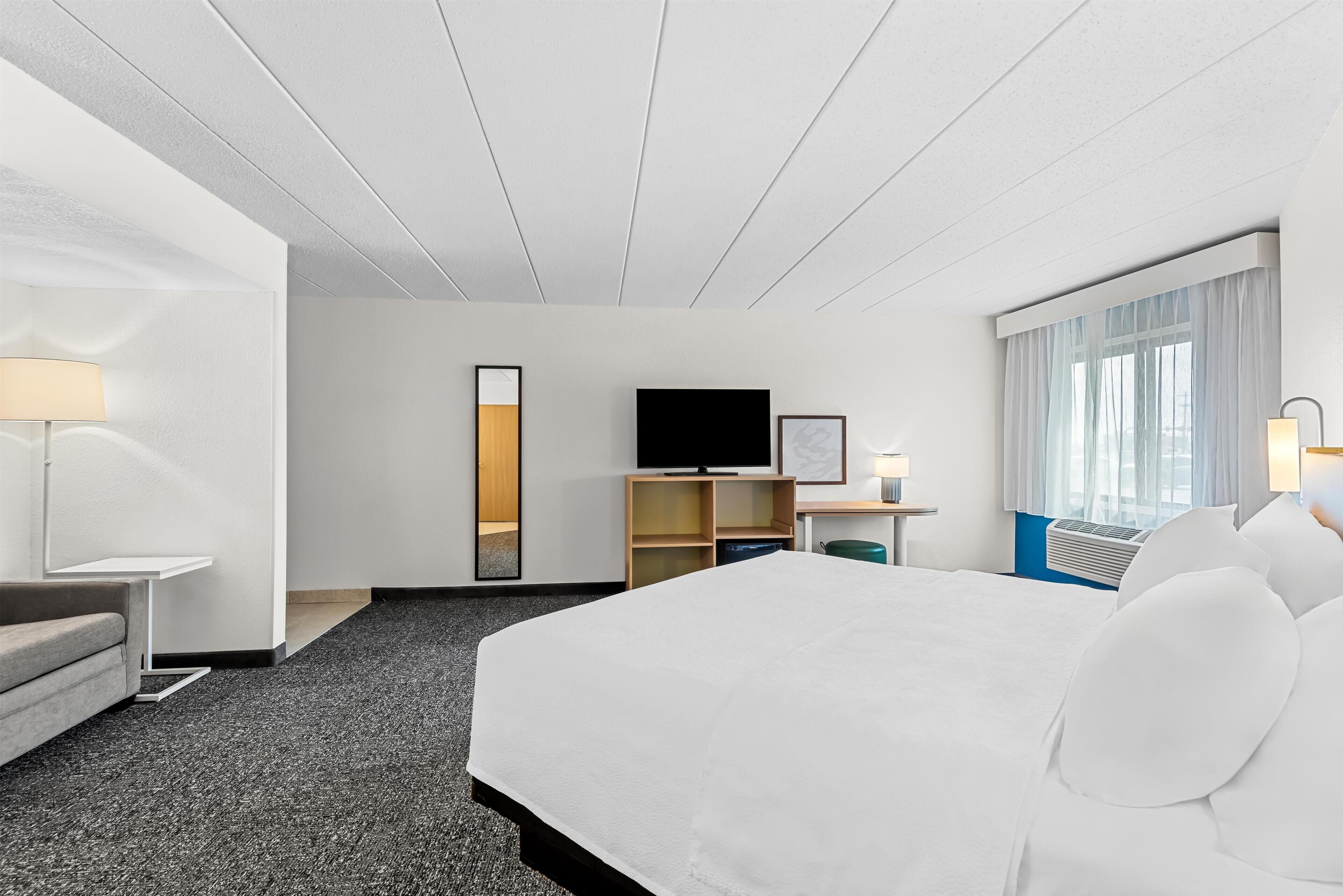 A hotel room with a large bed, a flat-screen TV, a desk with a lamp, a mirror, and a window with curtains.