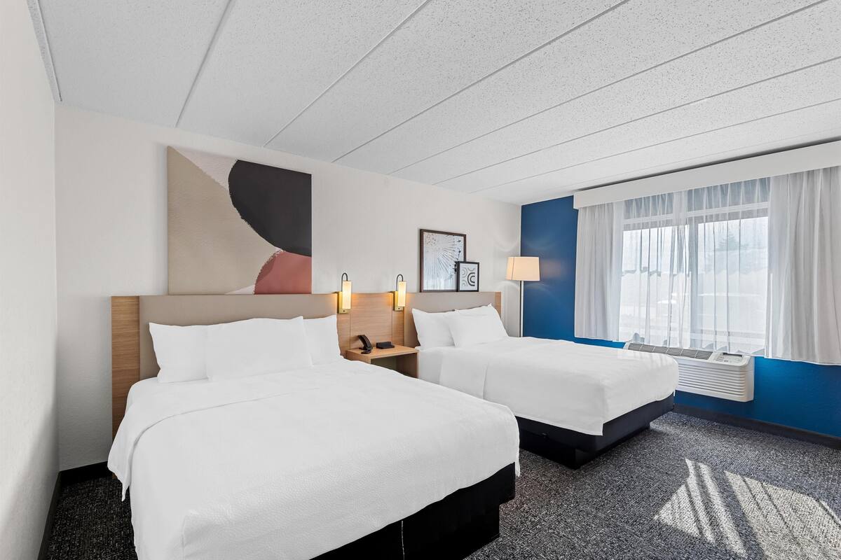 Habitación de hotel con dos camas, un cuadro abstracto grande, una ventana con cortinas translúcidas y un aire acondicionado de pared.