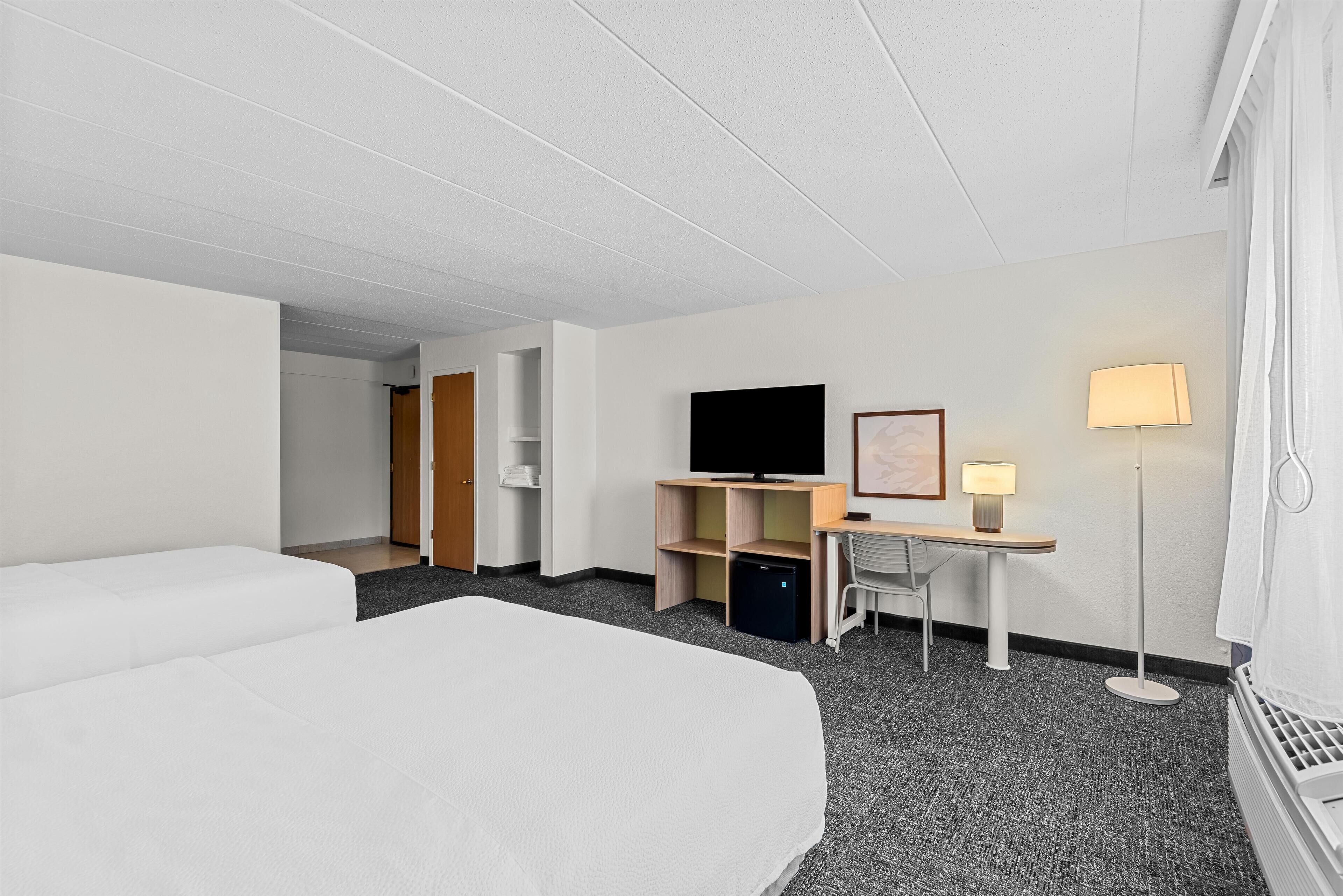 Hotellihuone, jossa on sänky, kirjoituspöytä lampun kanssa, televisio ja pieni oleskelualue.