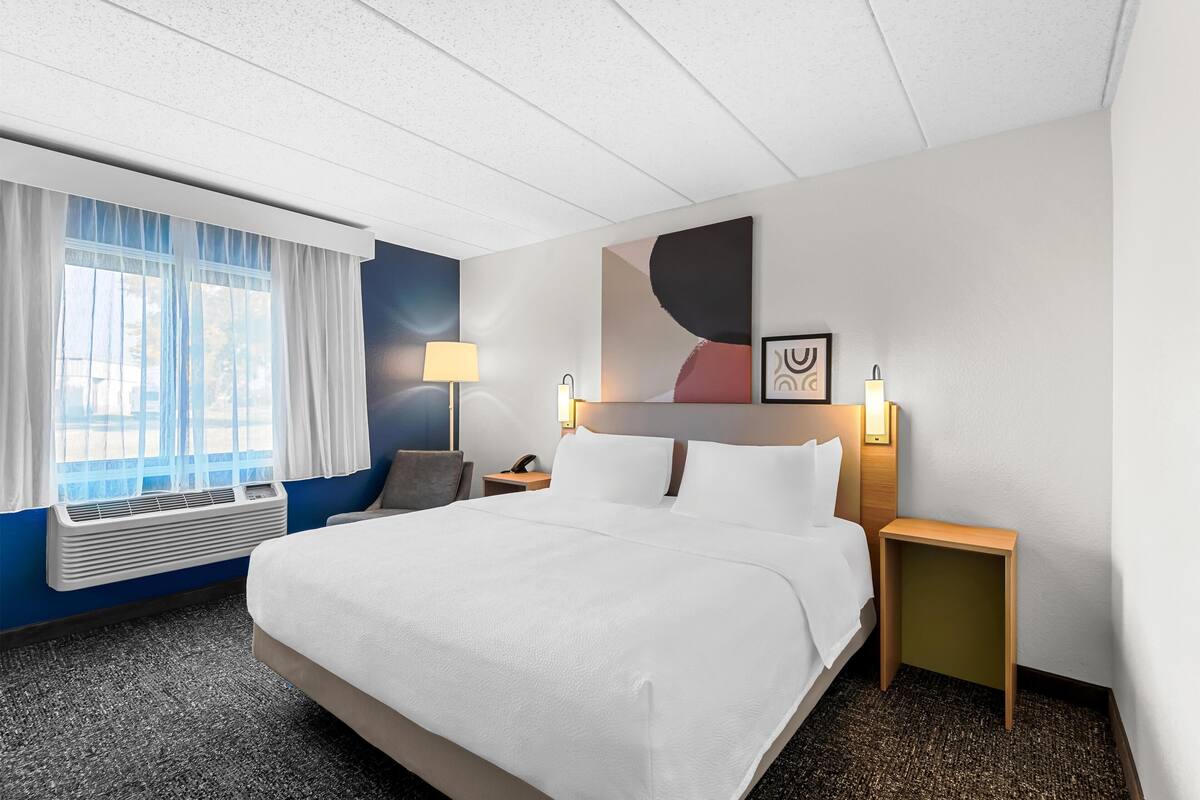 Una habitación de hotel con una cama grande, una mesita de noche con lámpara, aire acondicionado en la pared y una ventana con cortinas.