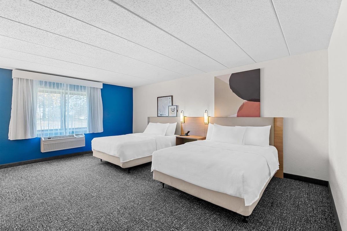 Habitación de hotel con dos camas, una ventana con cortinas, aire acondicionado de pared y una mesita de noche con lámpara.