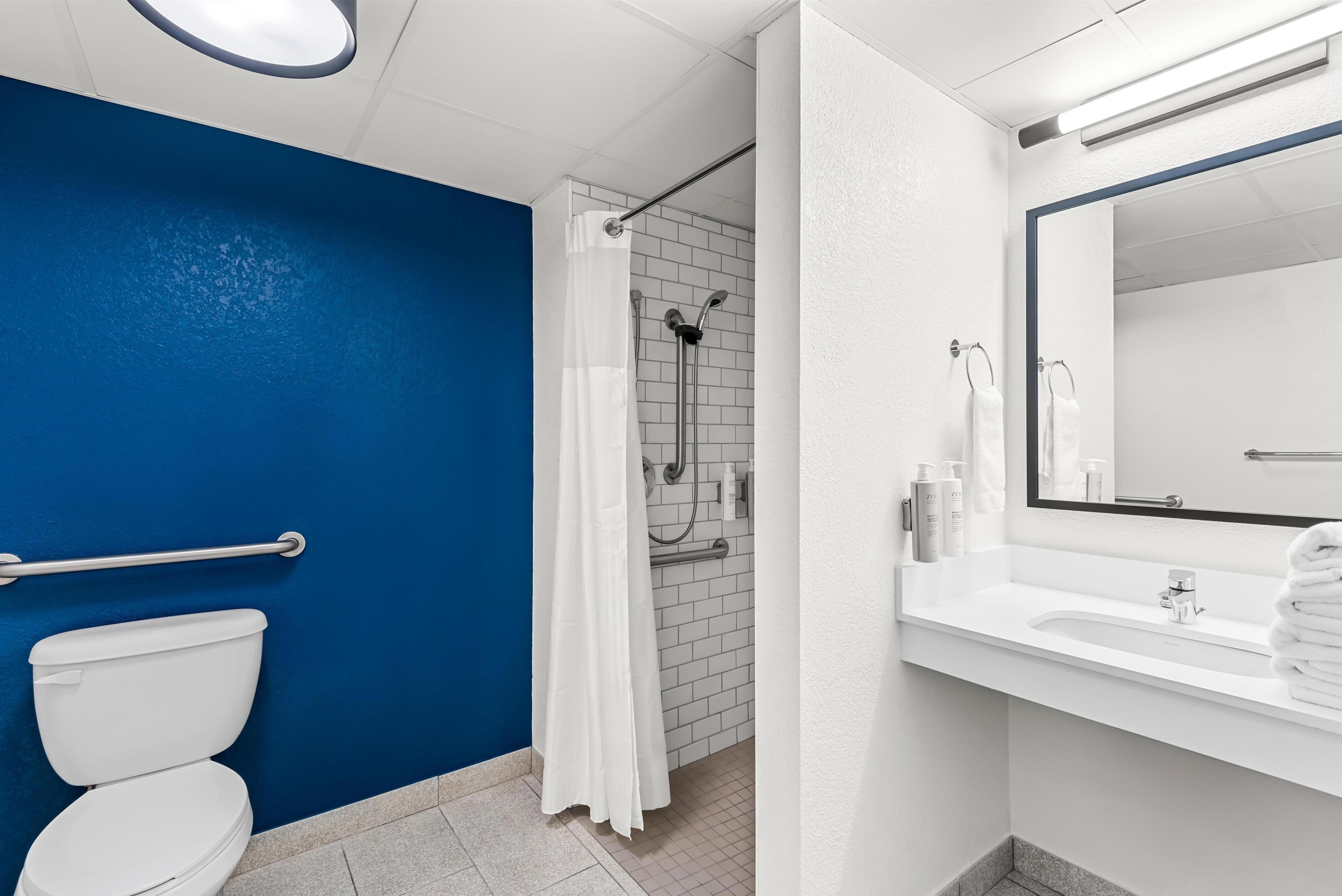 Un baño con pared azul, inodoro blanco, zona de ducha y lavabo con espejo.