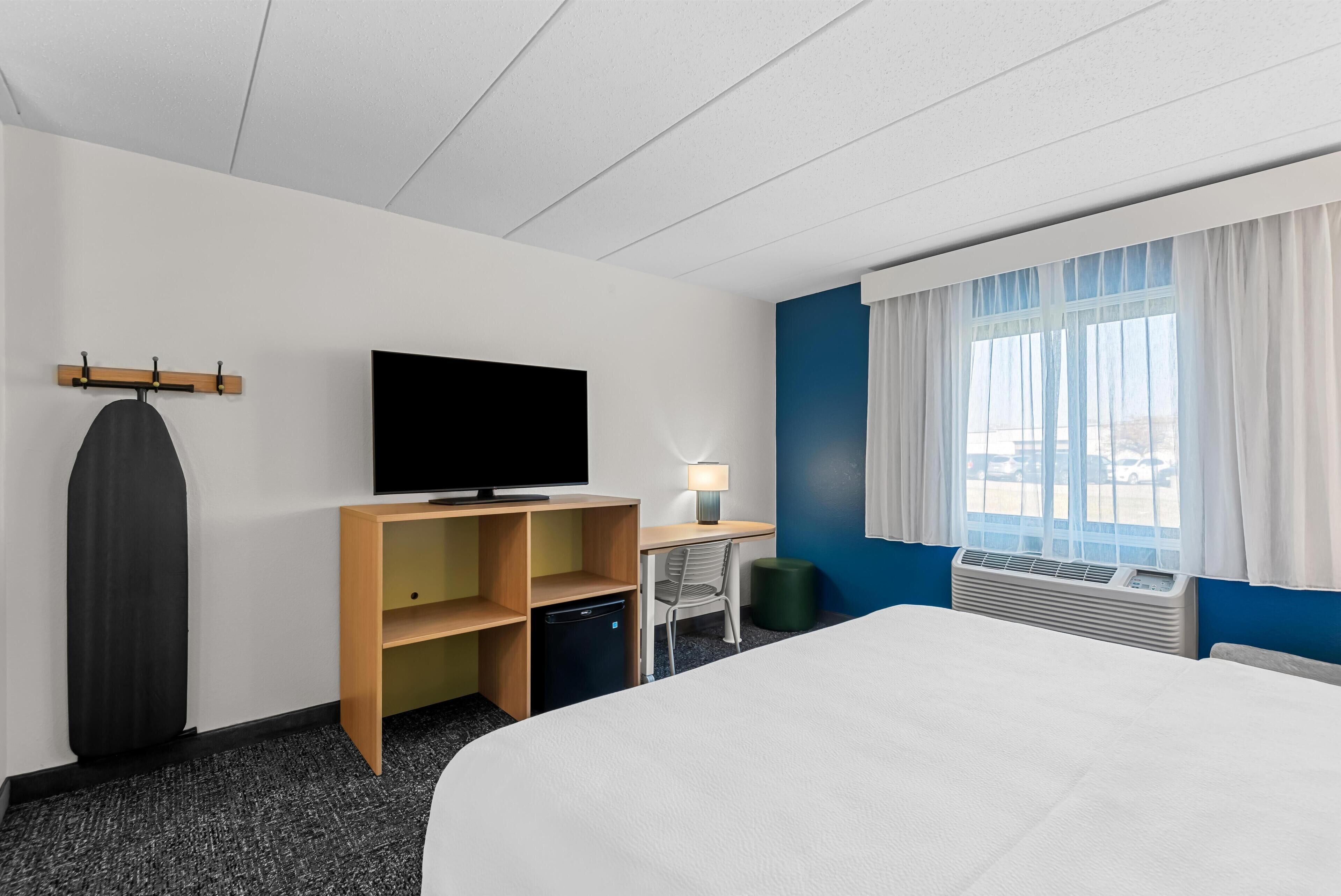 Habitación de hotel con cama, escritorio y silla, televisión, calefactor pequeño y ventana con cortinas.
