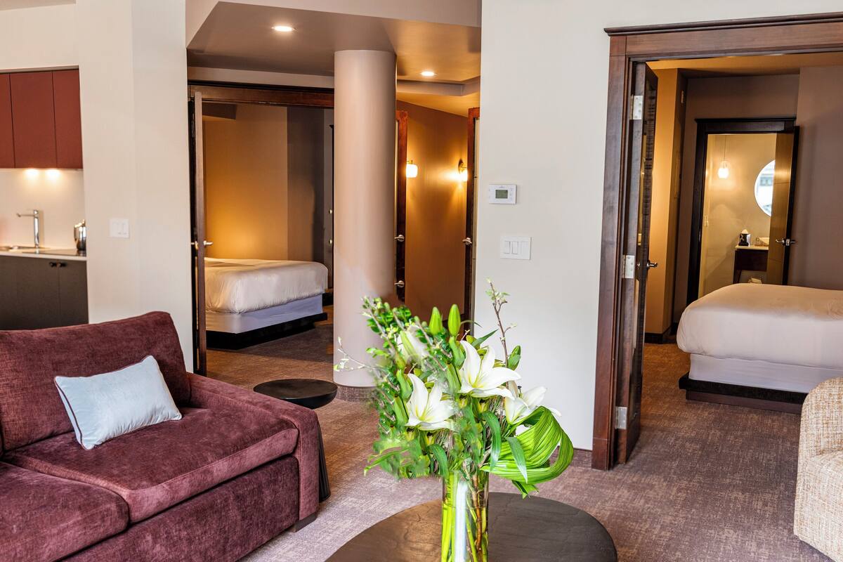 Une chambre d’hôtel avec un canapé, un lit, une petite table sur laquelle est posé un vase de fleurs, et une salle de bain visible à travers une porte ouverte.