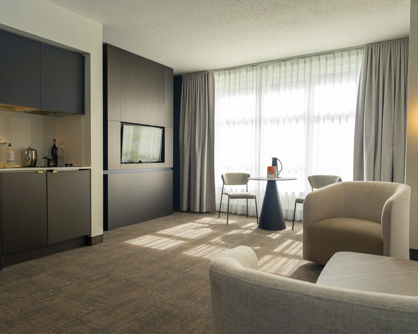 A hotel room with a kitchenette, a flat-screen TV, a dining area with two chairs, and a large window with curtains.