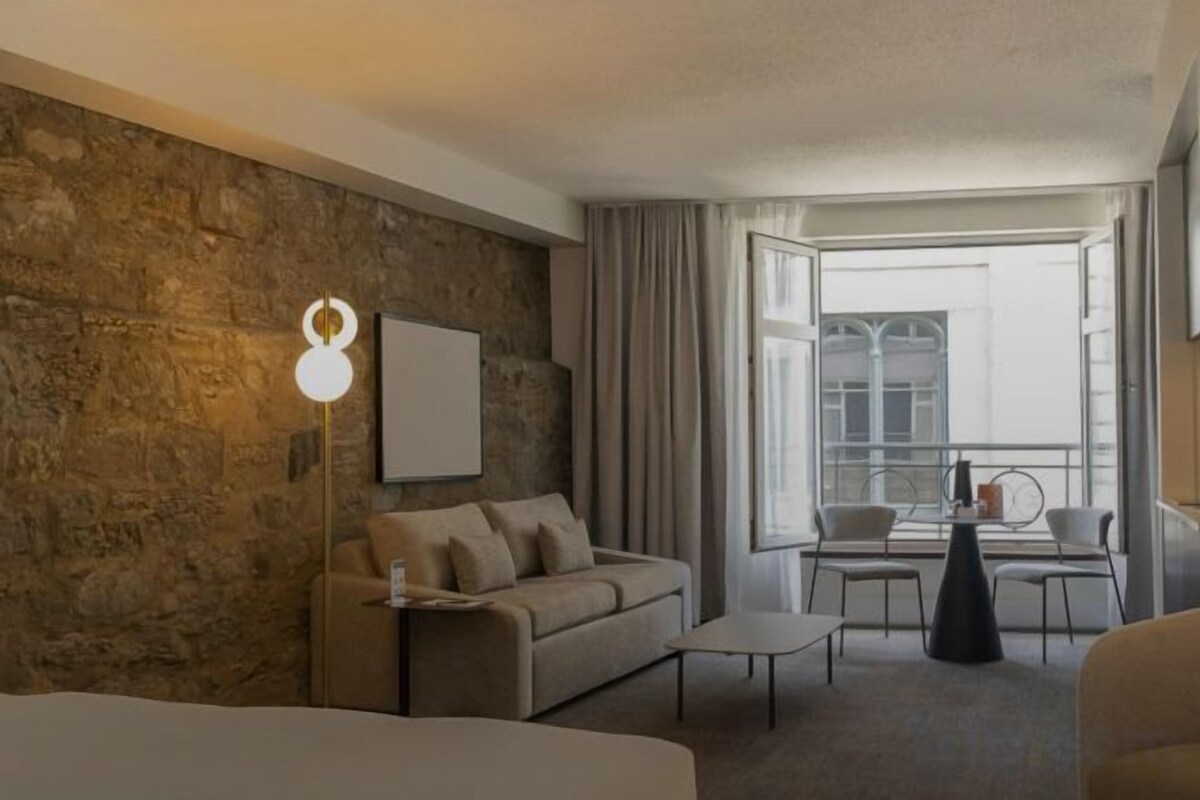A hotel room with a stone wall, a sofa, a coffee table, and a view of the city through large windows.