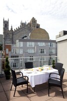 An outdoor dining area with a table set for two, surrounded by wicker chairs, overlooking a historic building with a large dome and spires.