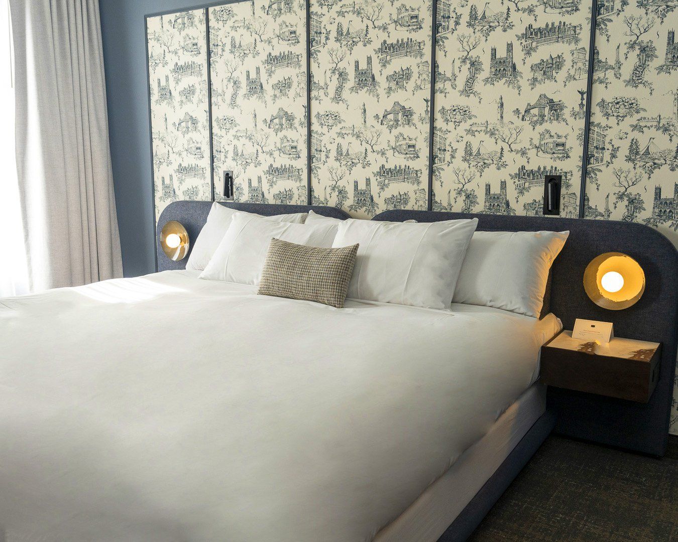 A hotel room with a large bed, white bedding, a patterned wallpaper, and two bedside lamps.