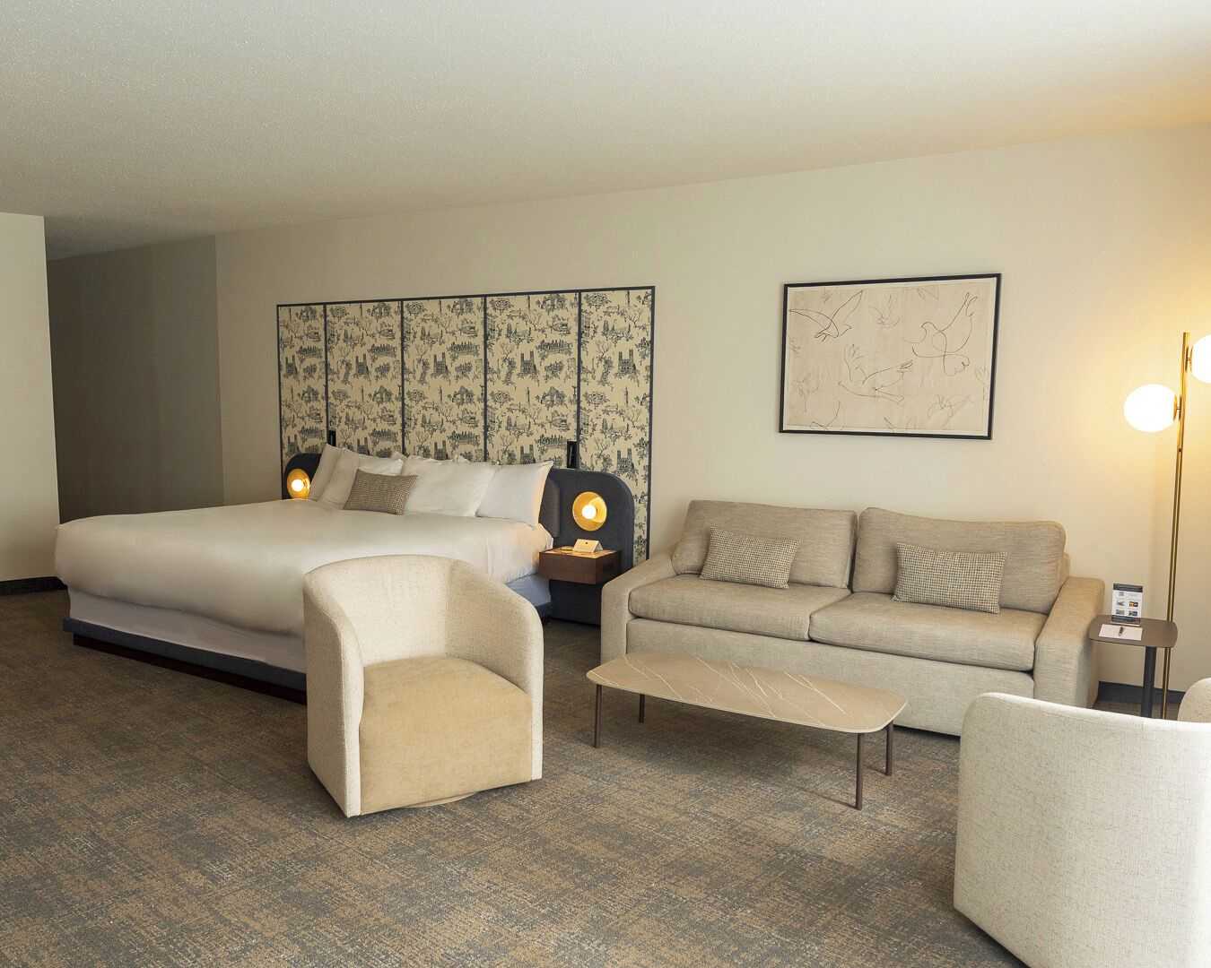 A hotel room with a large bed, a sofa, a chair, and a small table. There is a wall with a patterned wallpaper and a framed picture.