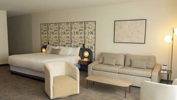 A hotel room with a large bed, a sofa, a chair, and a small table. There is a wall with a patterned wallpaper and a framed picture.