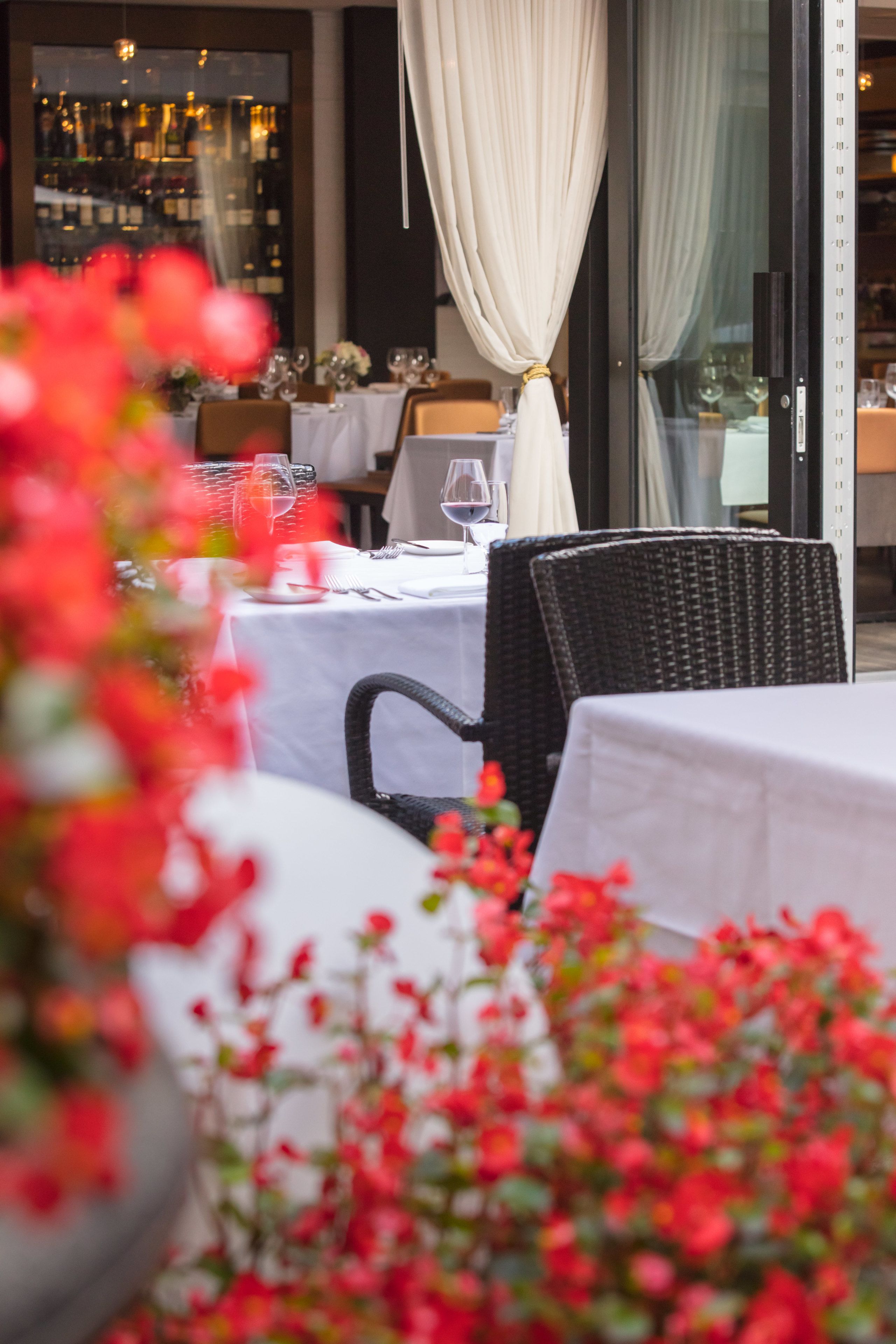 An outdoor dining area with tables set for two, featuring white tablecloths and black wicker chairs.