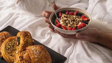Eine Person hält eine Schüssel mit Müsli, Früchten und Granola, daneben ein Teller mit Gebäck auf einem Bett.