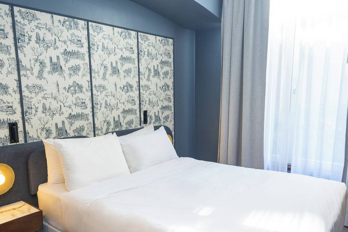 Une chambre d’hôtel avec un grand lit, une table de chevet avec une lampe et un mur recouvert d’un papier peint représentant un paysage urbain.