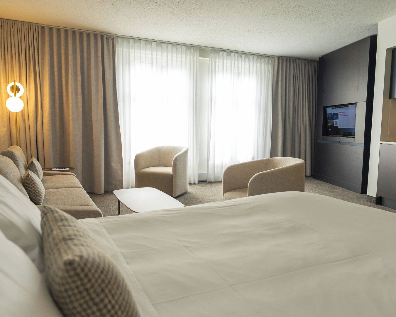 Ett modernt hotellrum med en stor säng, två fåtöljer, ett litet bord och en TV.