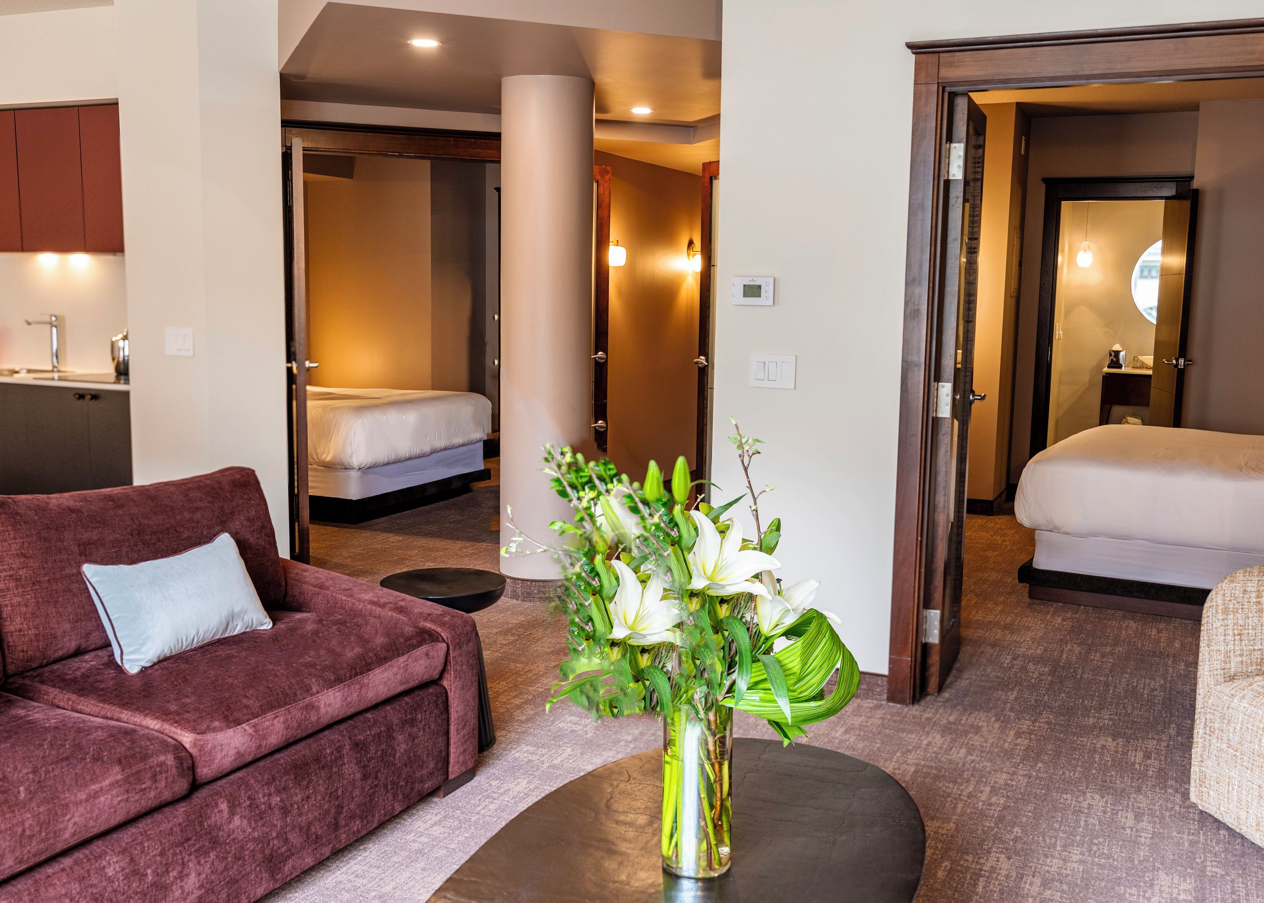 Una habitación de hotel con sofá, cama, una mesita con un jarrón de flores y un baño visible a través de una puerta abierta.