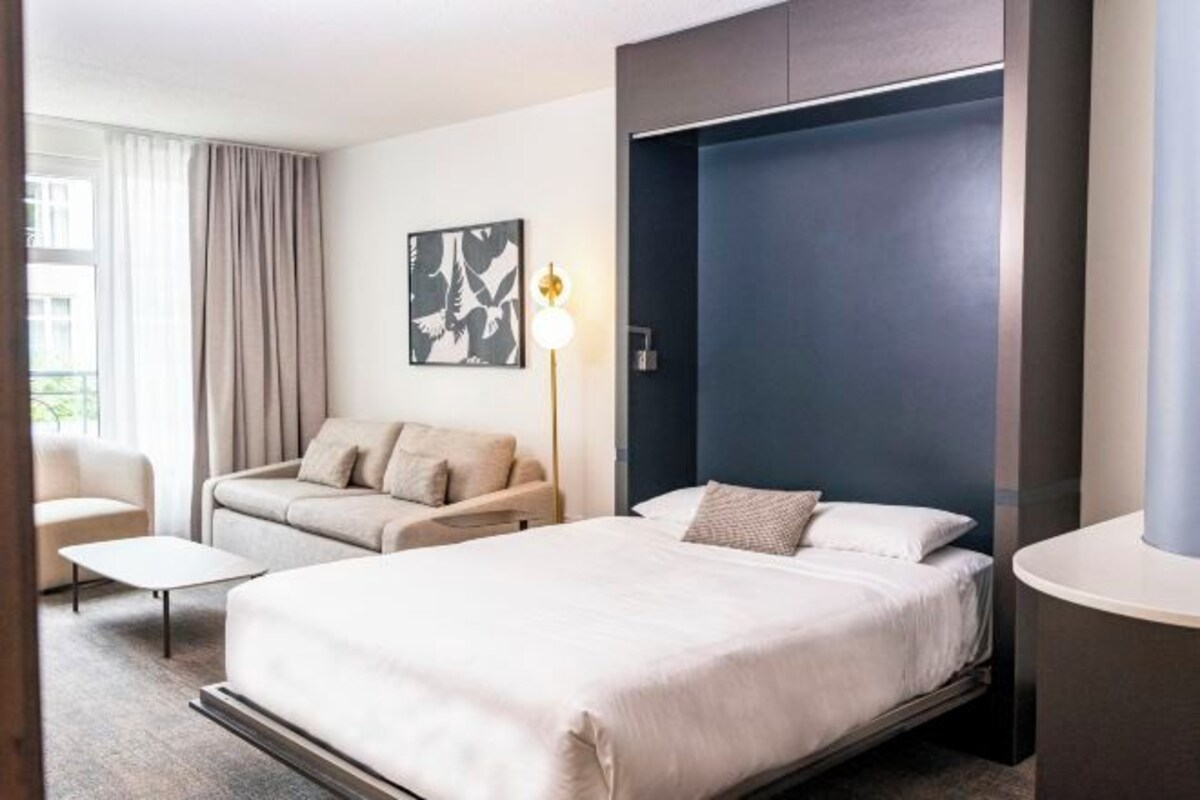 A hotel room with a bed, a sofa, a chair, a desk, a lamp, and a painting on the wall.