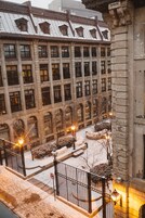 Une cour enneigée avec un bâtiment doté de grandes fenêtres et d’une façade en pierre.
