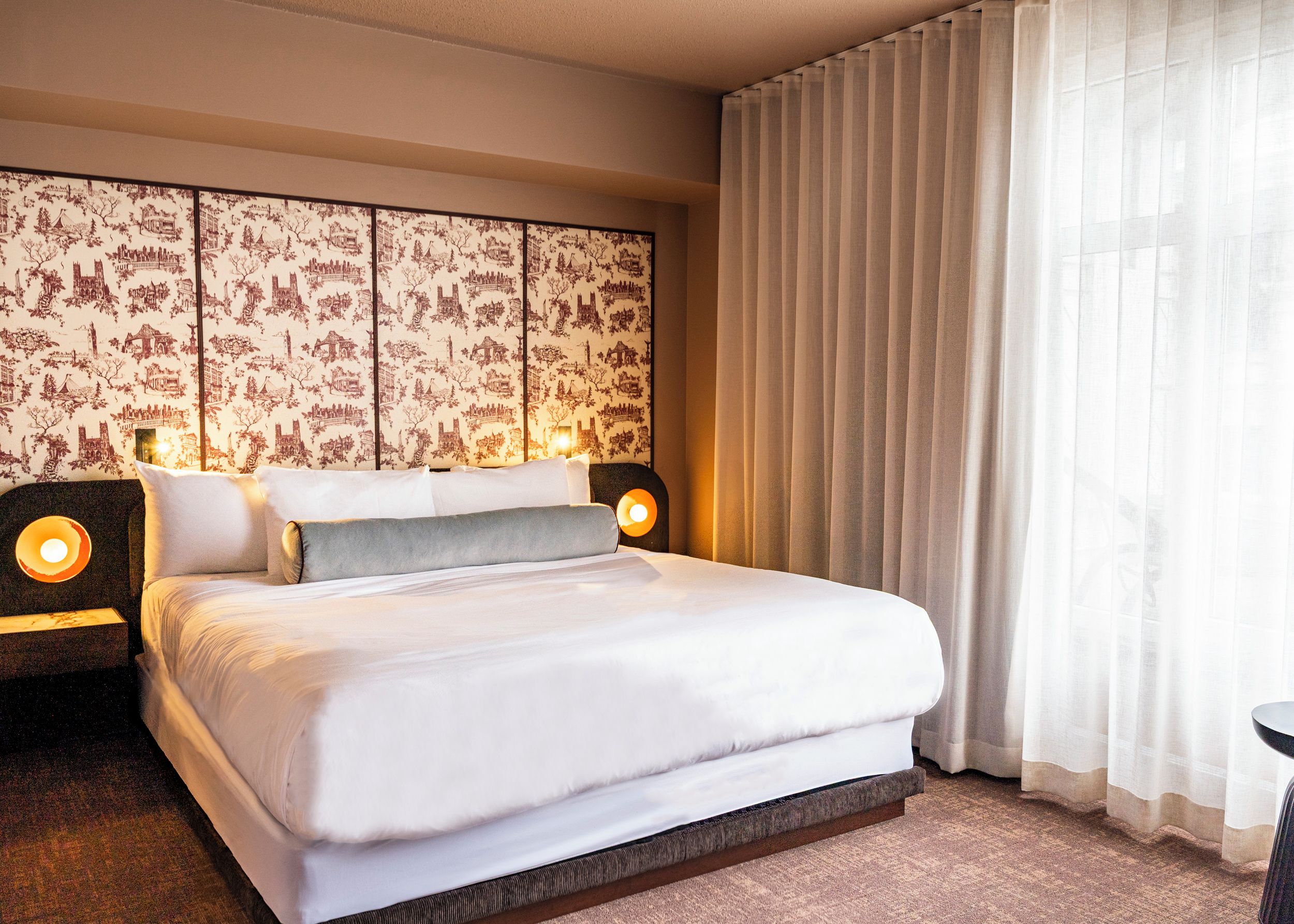 Une chambre d’hôtel avec un grand lit, deux lampes de chevet, un papier peint à motifs et des rideaux légers.