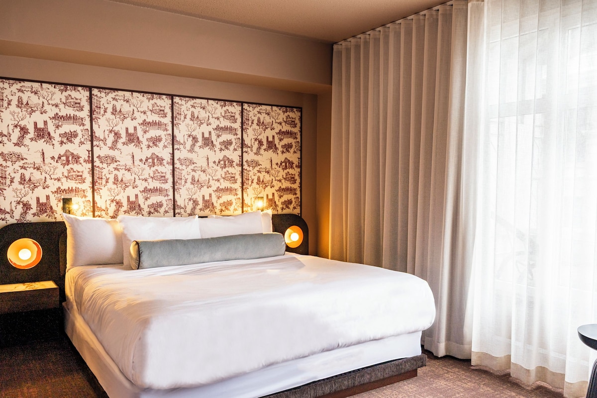 Une chambre d’hôtel avec un grand lit, deux lampes de chevet, un papier peint à motifs et des rideaux légers.