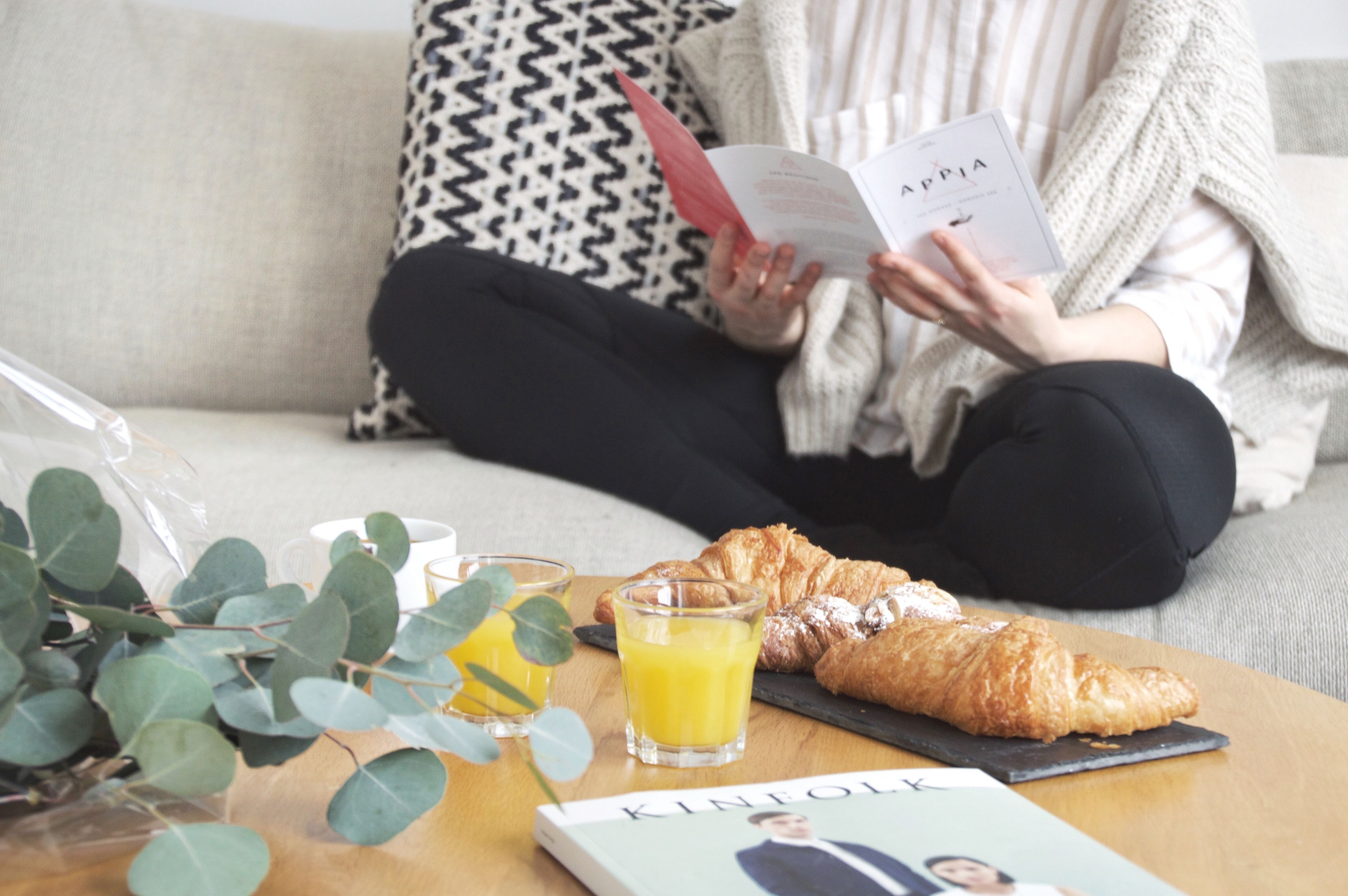 A person sitting on a couch with a book, a croissant, and a glass of orange juice on a table.