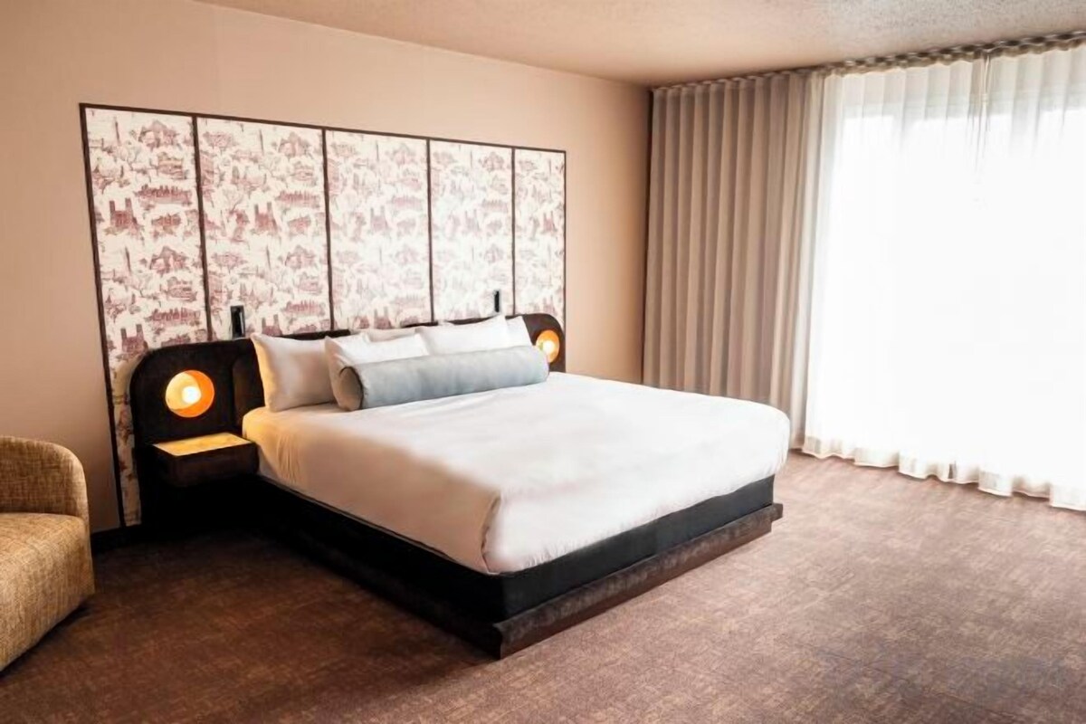 A hotel room with a large bed, a headboard with a decorative panel, a nightstand with a lamp, and a window with curtains.