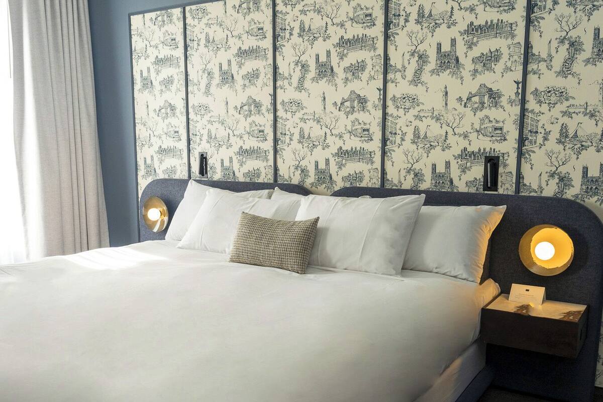 Un lit bien fait, avec du linge de lit blanc et une tête de lit à motifs. Il y a deux tables de chevet avec des lampes.