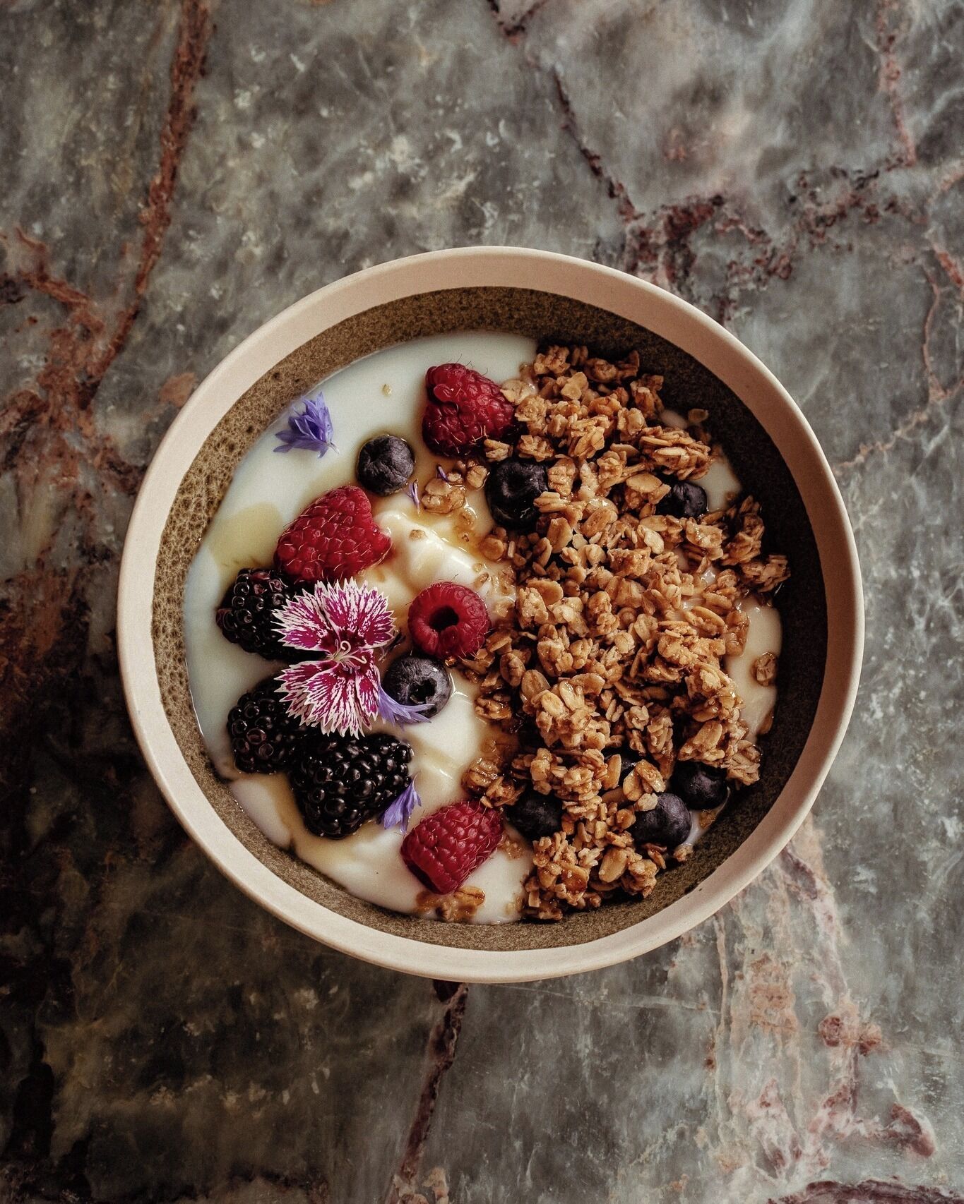 Un tazón de yogur con granola y bayas sobre una superficie de mármol.