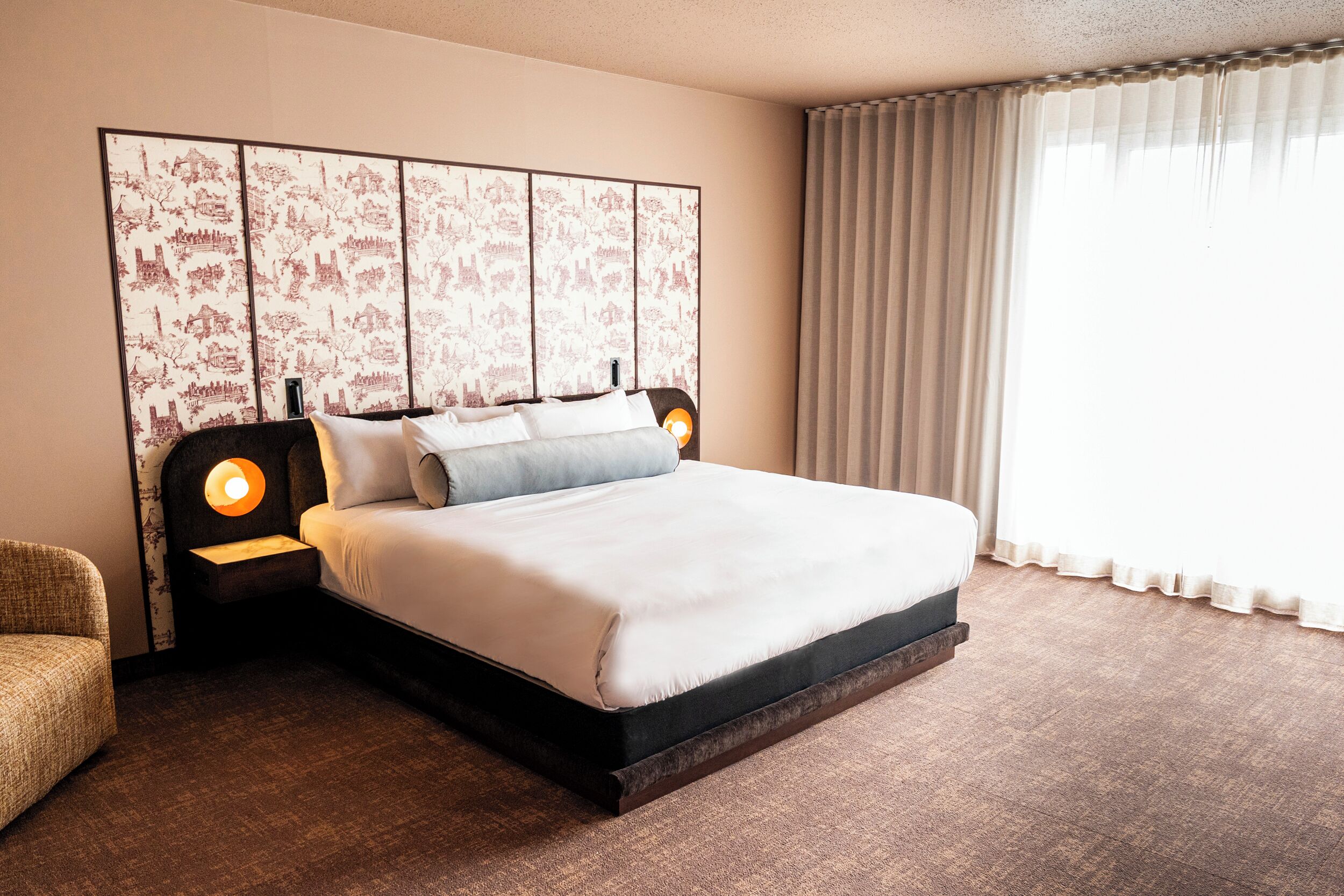 Quarto de hotel bem organizado, com uma cama grande, cabeceira com painel decorativo, mesa de cabeceira com luminária e janela com cortinas.