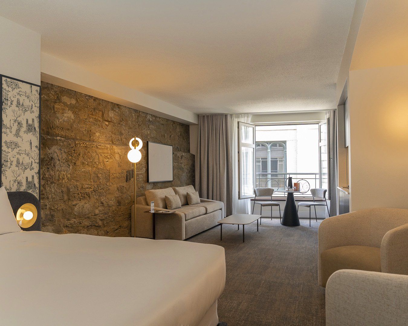 Une chambre d’hôtel avec un mur en pierre, un lit, un canapé, une table à manger avec des chaises, et une vue sur la ville grâce à de grandes fenêtres.