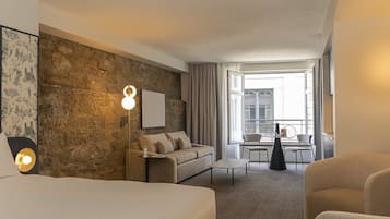 Habitación de hotel con pared de piedra, cama, sofá, mesa de comedor con sillas y vistas a la ciudad a través de amplias ventanas.