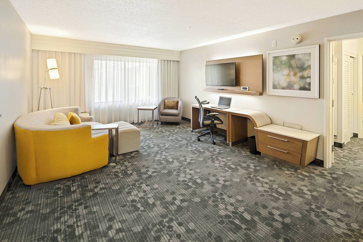 A hotel room with a yellow sofa, a desk with a computer, a TV, and a large window with curtains.