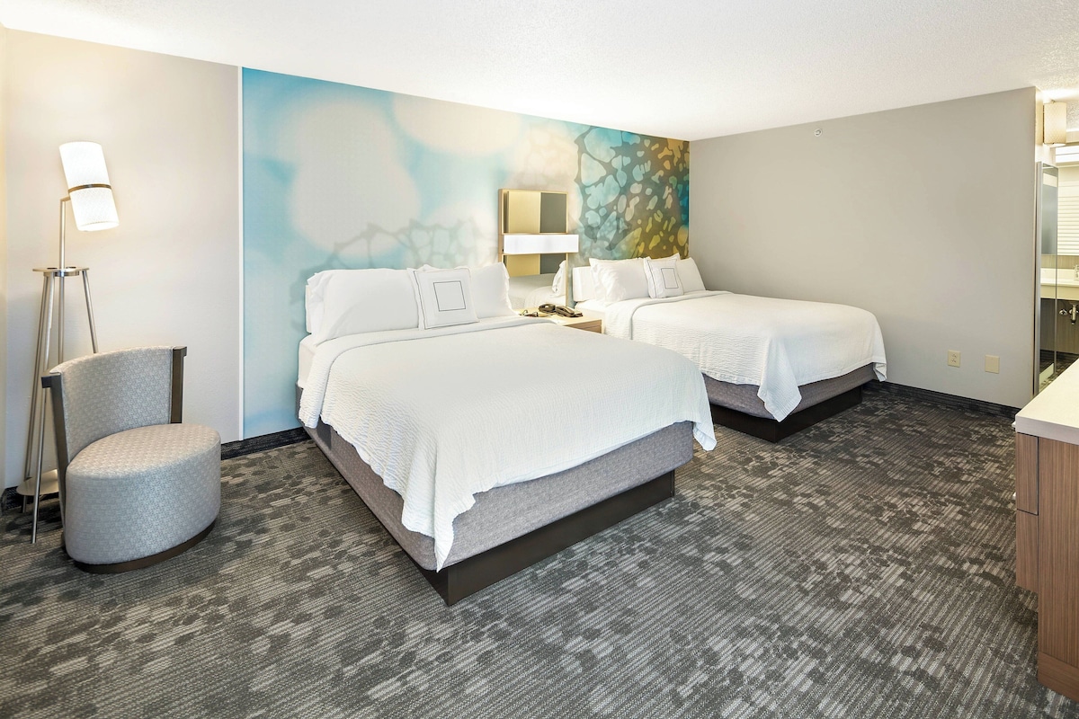 Ein Hotelzimmer mit zwei Betten, einem Sessel, einem Schreibtisch und einem Spiegel.