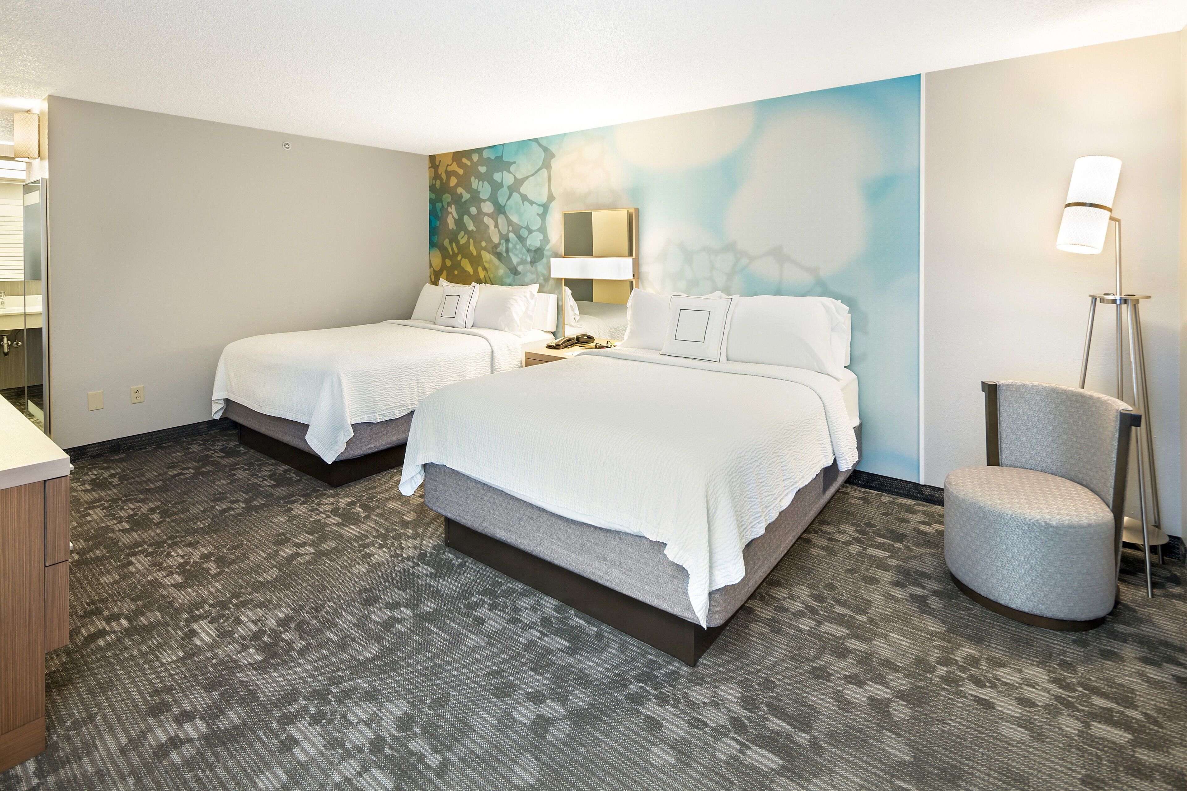 A hotel room with two beds, a desk, a chair, a lamp, and a painting on the wall.