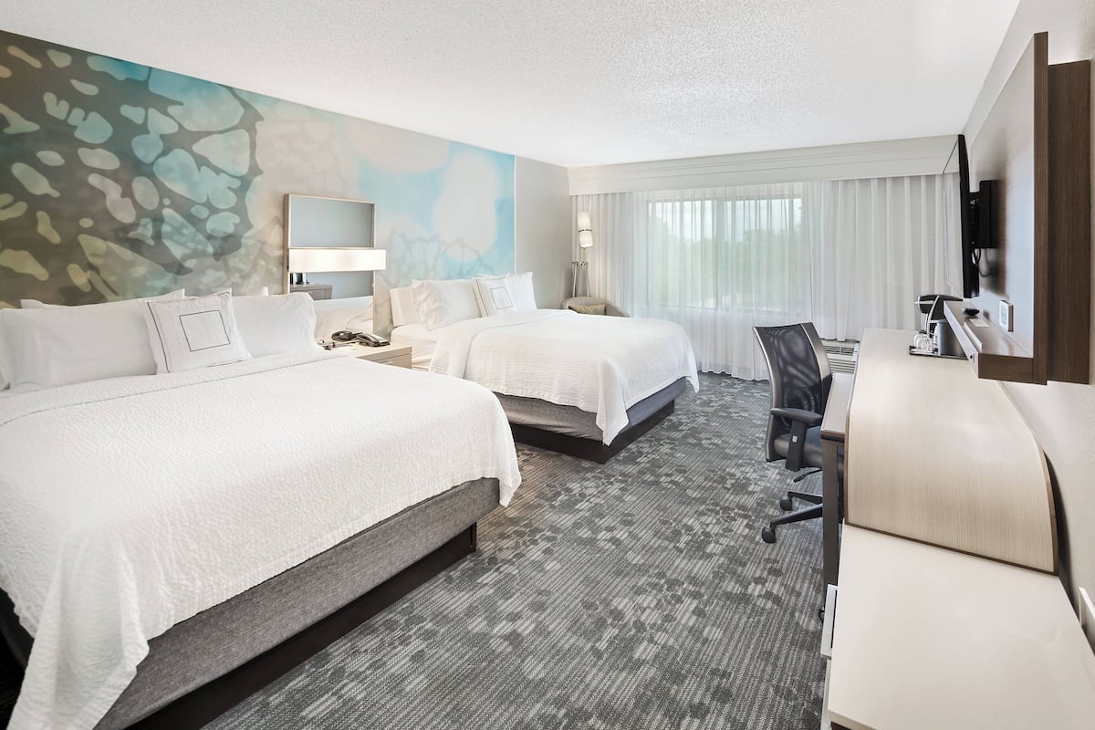 A hotel room with two beds, a desk, a chair, a TV, and a large window with curtains.