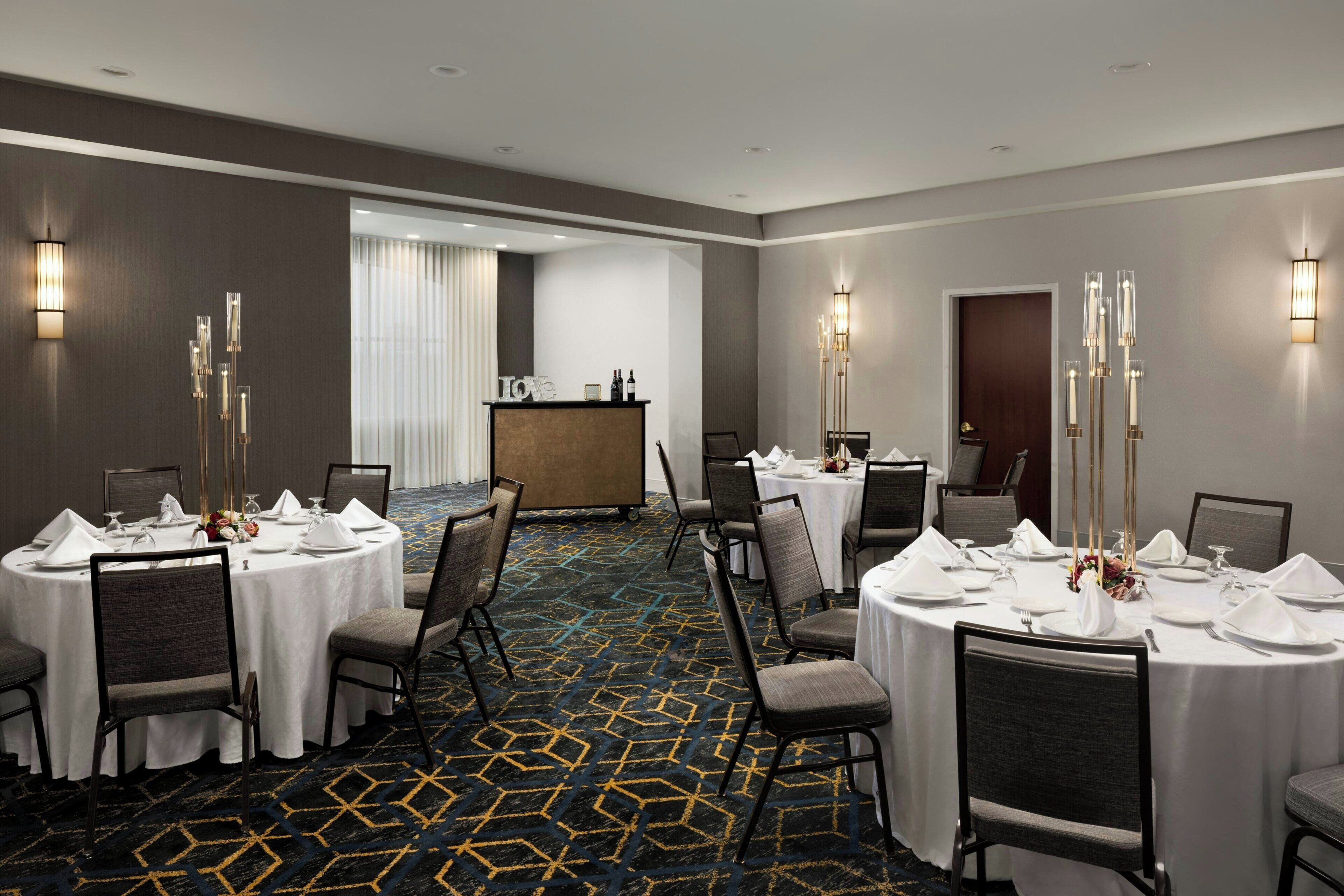 Un comedor de hotel con mesas redondas listas para el servicio, una recepción y una alfombra estampada.
