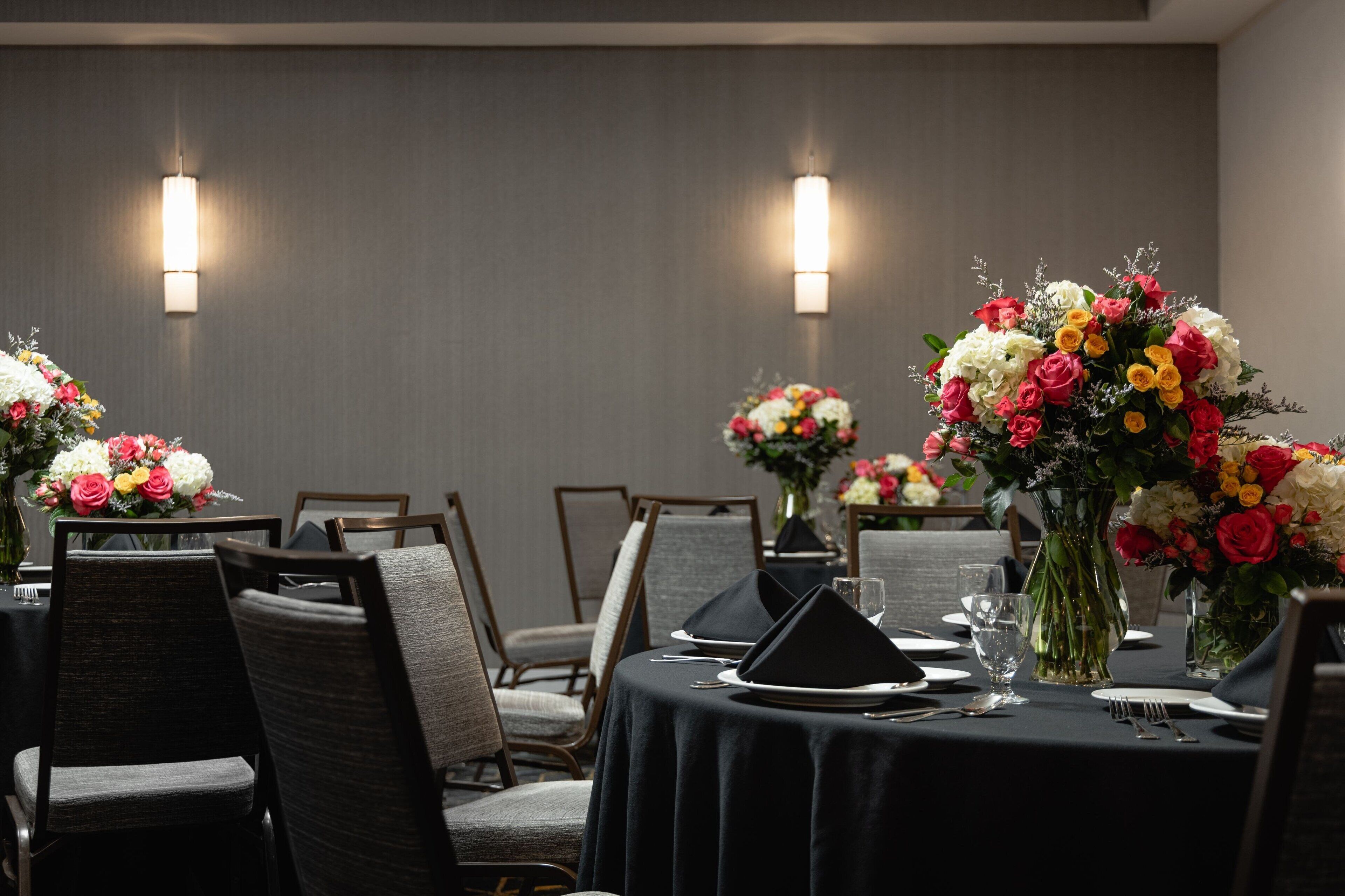 A dining area with tables set for four, each with a black tablecloth, white plates, and black napkins. There are floral centerpieces on each table, and the walls are adorned with wall sconces.