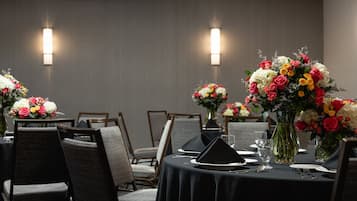 Comedor con mesas dispuestas para cuatro, cada una con mantel negro, platos blancos y servilletas negras. Los centros de mesa son arreglos florales y las paredes están decoradas con apliques de pared.