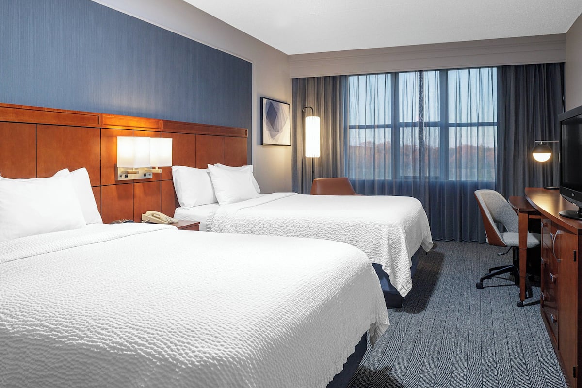 A hotel room with two beds, a desk, a chair, a TV, and a large window with curtains.