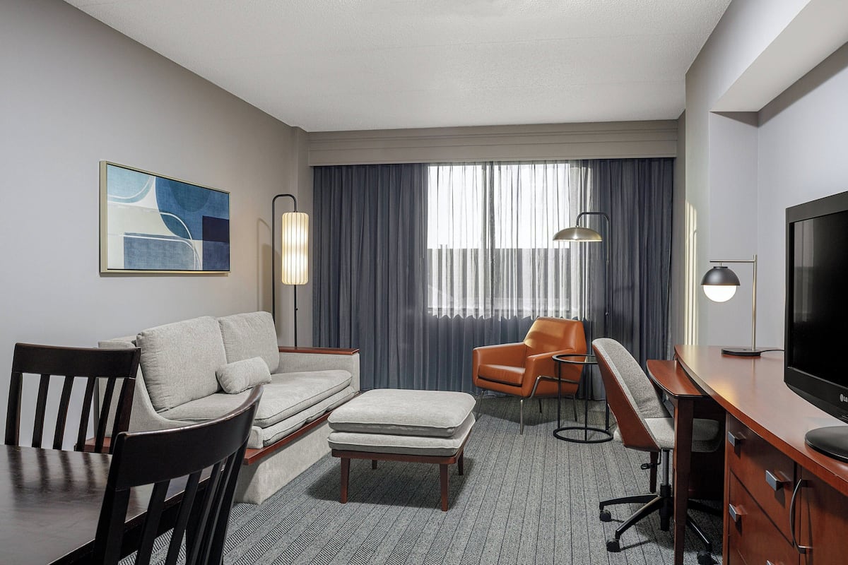 A hotel room with a sofa, armchair, ottoman, desk, and TV.