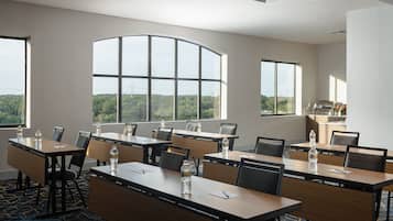 Un salón de conferencias con varias mesas y sillas, ventanas amplias con vistas a zonas verdes, y una cocina visible a través de las ventanas.