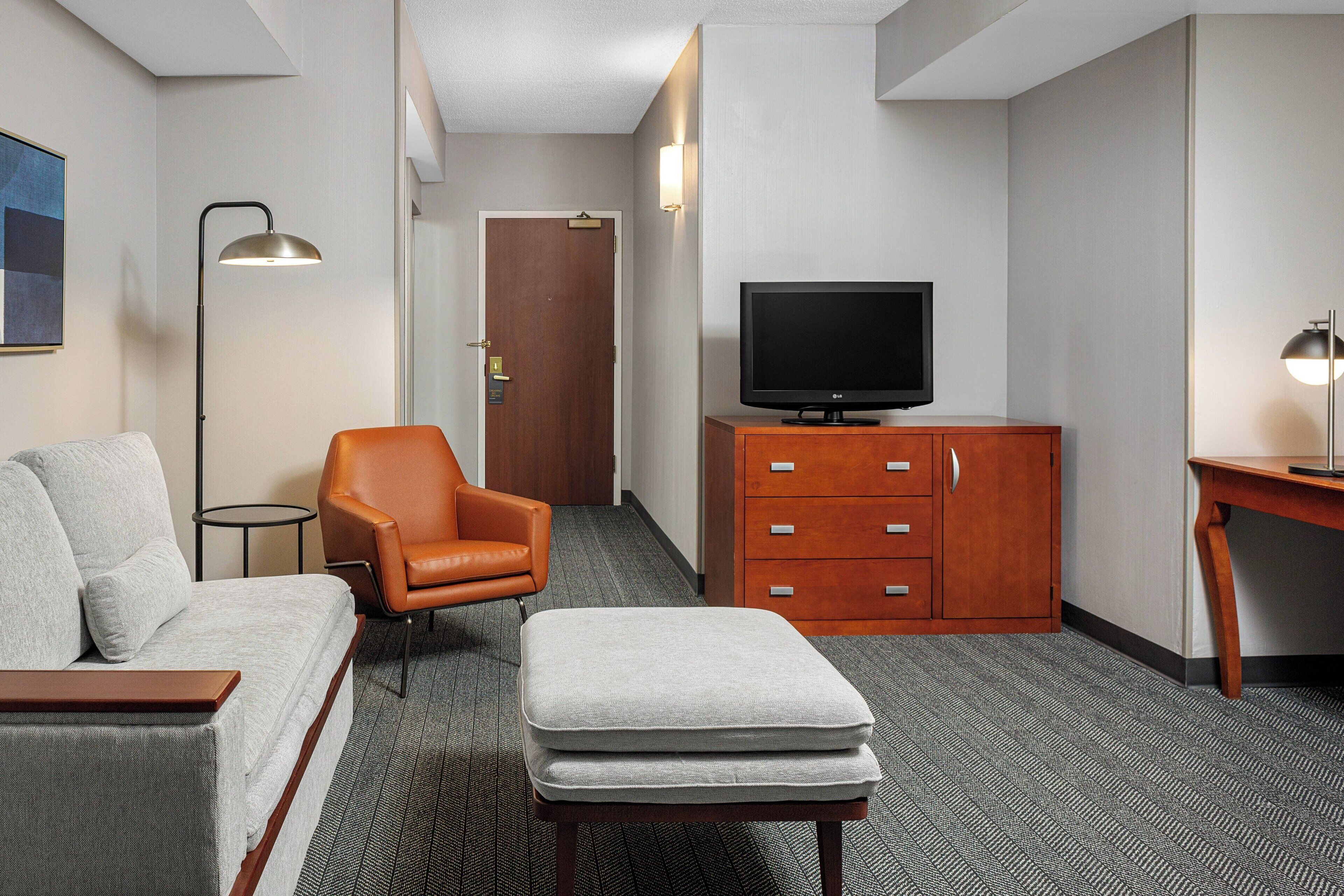 Une chambre d’hôtel comprenant un canapé, un pouf, un meuble en bois avec un téléviseur et un bureau avec une lampe.