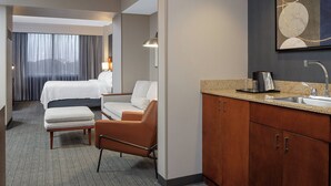 Habitación de hotel con cama, sofá, una mesita, sillón, kitchenette y un amplio ventanal con cortinas.