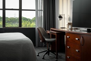 Habitación de hotel con cama, escritorio con silla, televisión y vista a zonas verdes a través del ventanal.