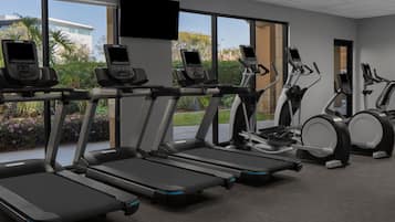 Un gimnasio con cintas de correr y máquinas elípticas, amplias ventanas con vistas a zonas verdes y edificios, y un televisor de pantalla plana montado en la pared.