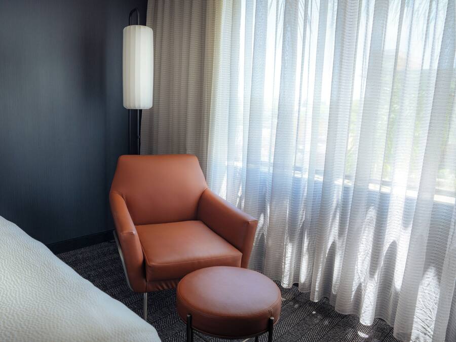 A hotel room with a bed, a brown armchair, a round ottoman, a floor lamp, and sheer curtains.