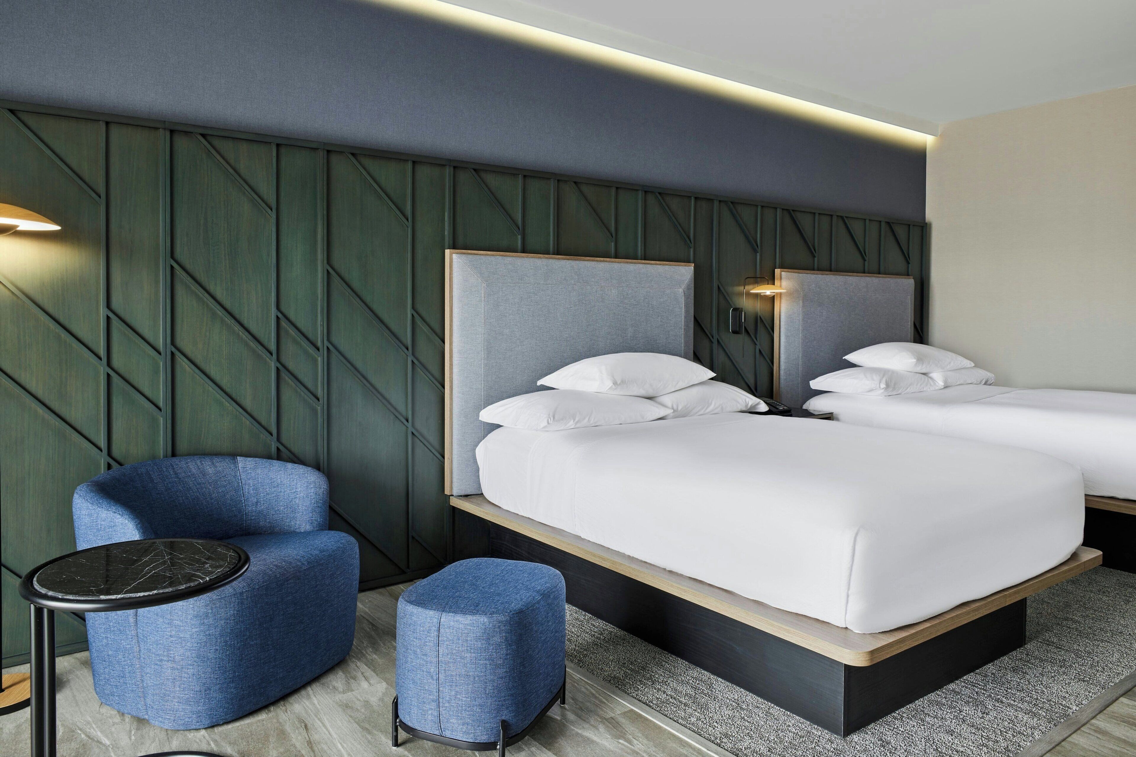 A modern hotel room with two beds, a blue armchair, a small ottoman, and a round table.